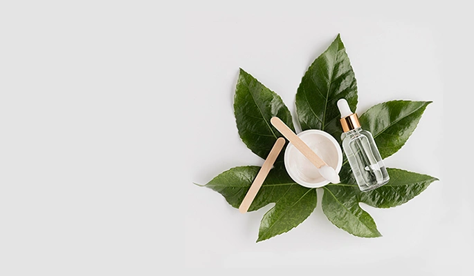 Pot de crème avec spatule en bois posé parmi des feuilles vertes brillantes sur fond beige clair